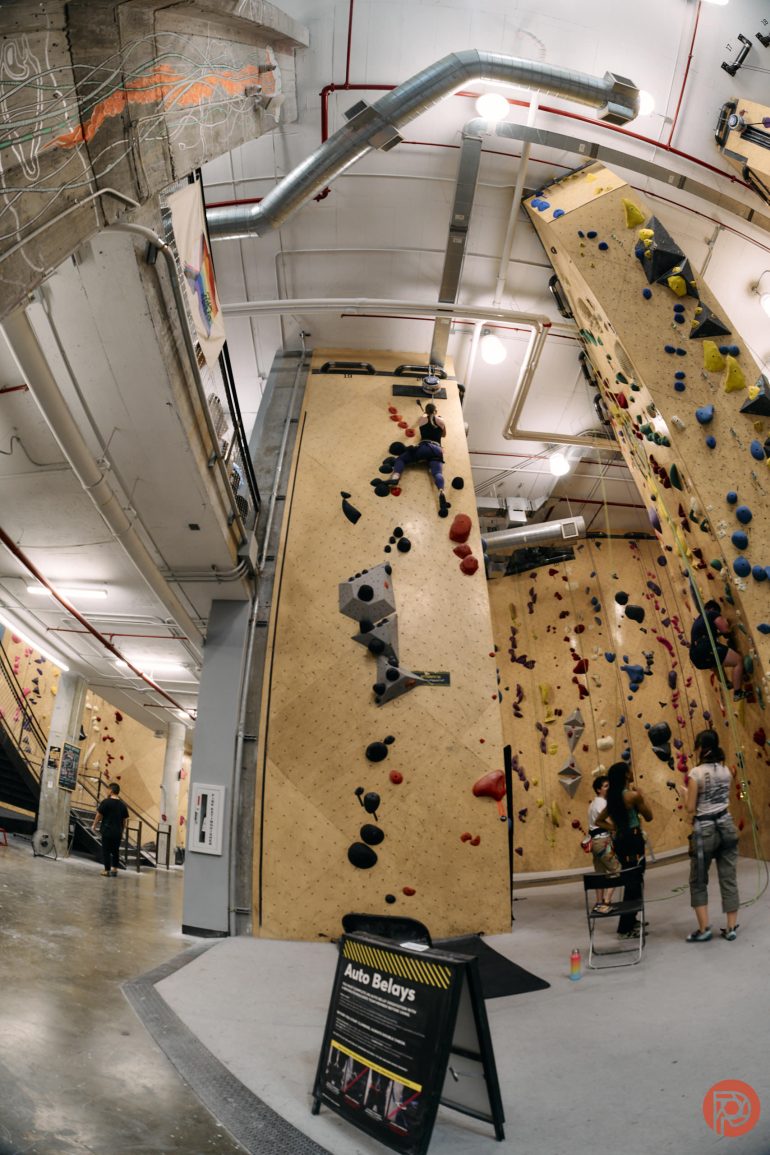 People indoor rock climbing on tall walls, with others watching or preparing at the bottom in a gym.