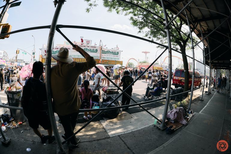 People watch a crowded outdoor event near Nathans Famous at Coney Island, seen through metal scaffolding.