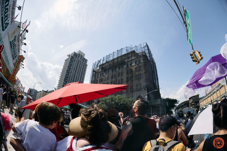 A crowd gathers under umbrellas on a sunny day near a building covered in scaffolding.