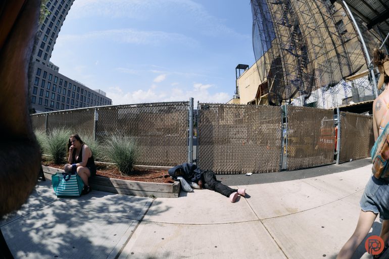 A person lies on a sidewalk bench while another sits nearby, next to a construction site and chain-link fence.