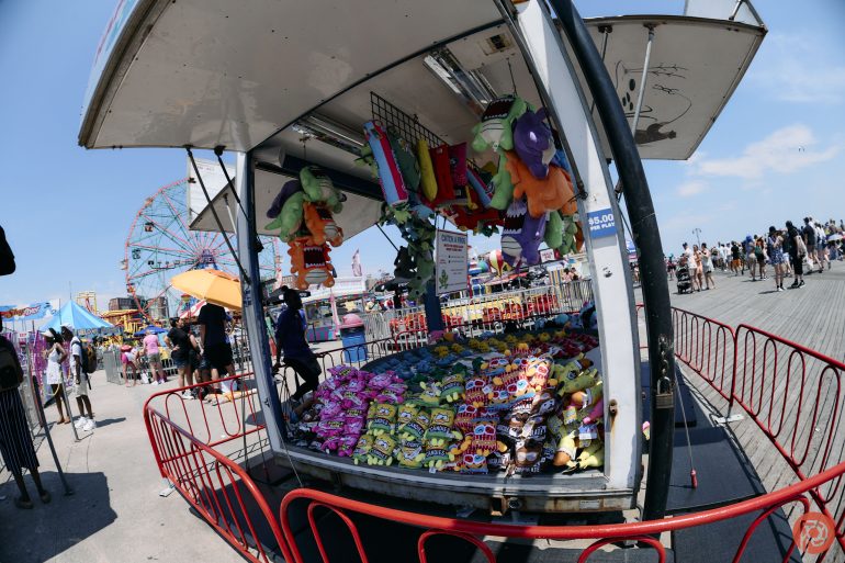 Carnival game booth with stuffed prizes, snacks, and people walking on a sunny boardwalk near a Ferris wheel.