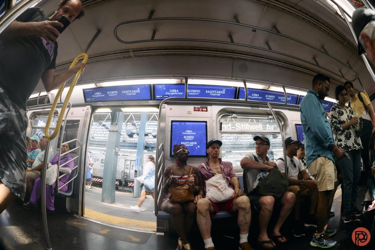 People sit and stand inside a subway train with doors open at a station; some are looking at phones.