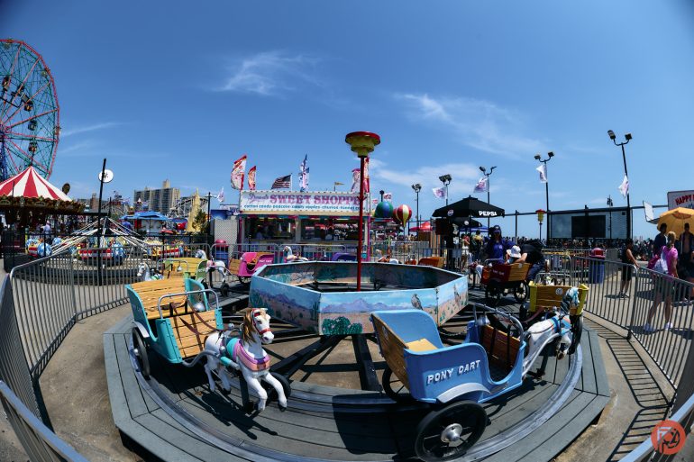 Children’s carnival ride with horse and cart seats at an amusement park, under a bright blue sky.