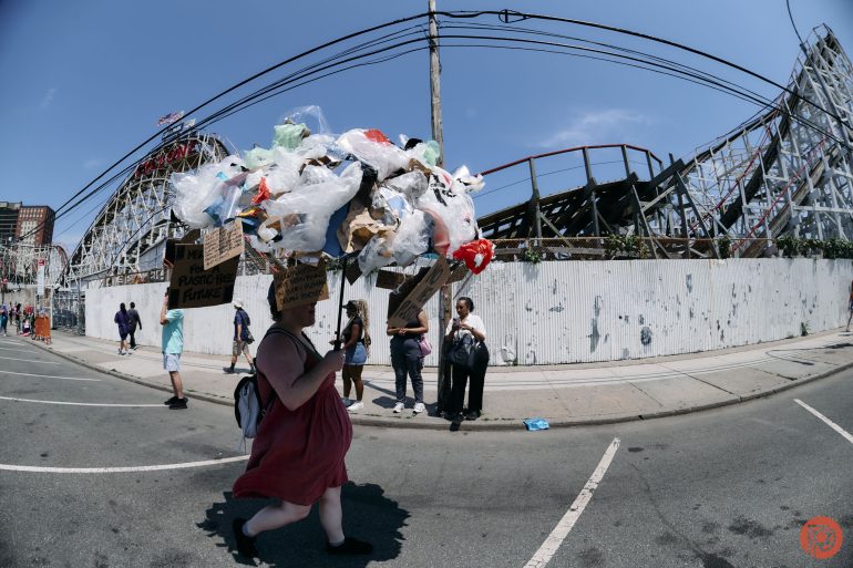 A person with a large hat made of plastic bags walks past a roller coaster, holding protest signs.