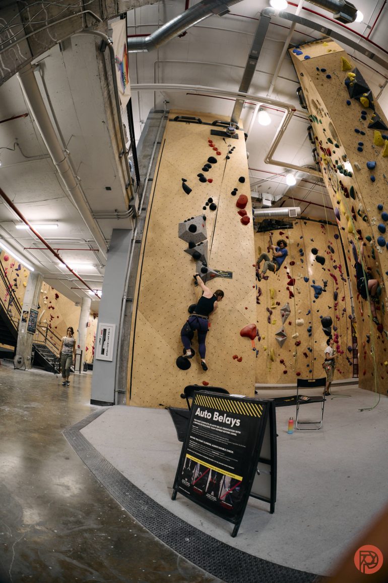 Person climbing an indoor rock wall; a sign about auto belays is visible in the foreground.