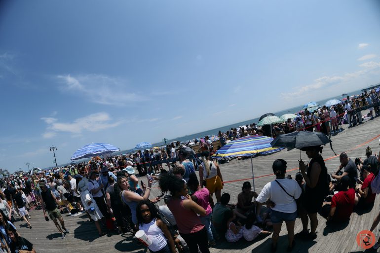 Large crowd gathered on a sunny boardwalk, many using umbrellas for shade, with the ocean in the background.