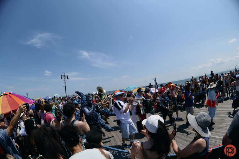 A lively parade with musicians and a blue sea creature costume on a sunny boardwalk surrounded by a crowd.