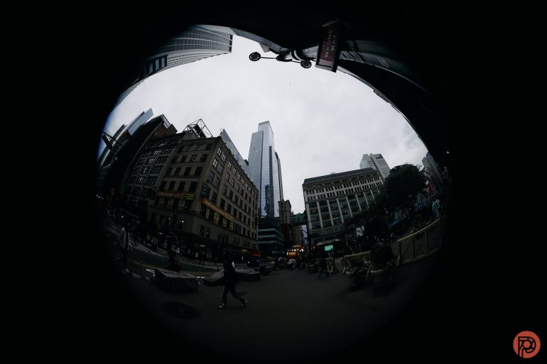 A fisheye lens view of a city street with tall buildings and people walking under a cloudy sky.