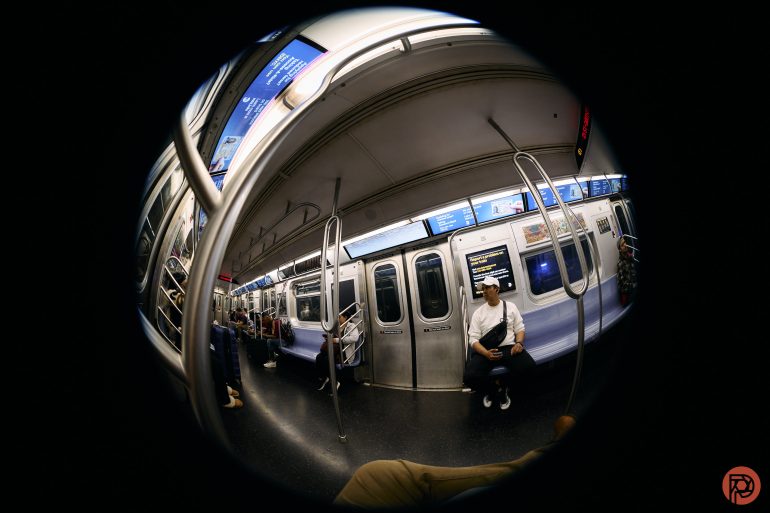 Fisheye view of people sitting in a subway car, with empty seats and overhead ads visible.