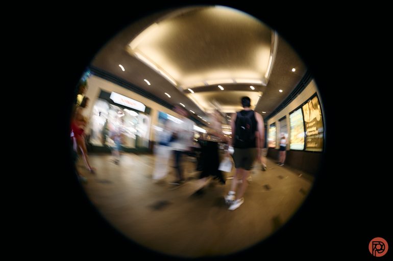 Fisheye view of people walking in a brightly lit mall hallway with blurred motion.