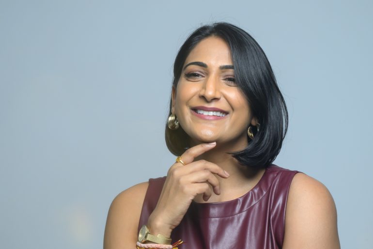 Woman with short black hair, smiling and resting her chin on her hand, wearing a sleeveless maroon top.