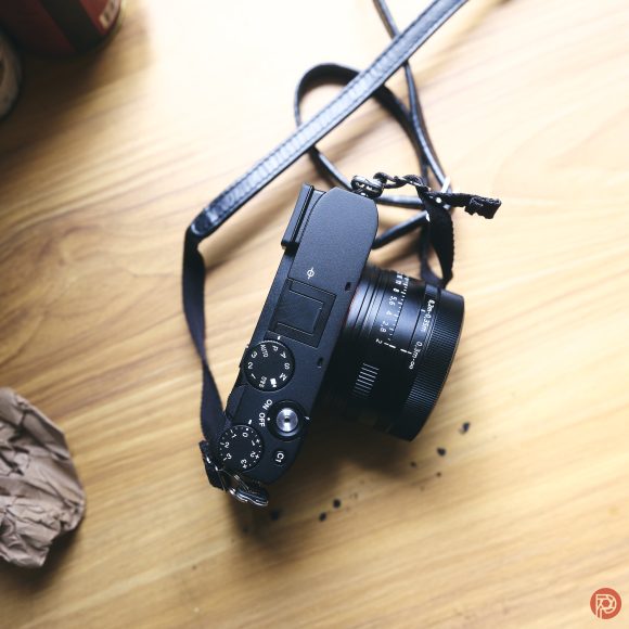 Black camera with a strap on a wooden surface, viewed from above, with some small dark specks nearby.
