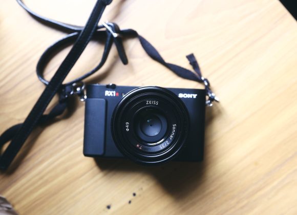 A black Sony RX1 camera with a Zeiss lens sits on a light wooden surface with its strap attached.