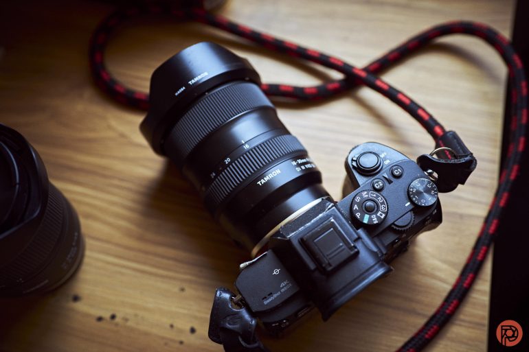 A black Sony camera with a Tamron lens and a red-black strap rests on a wooden surface.