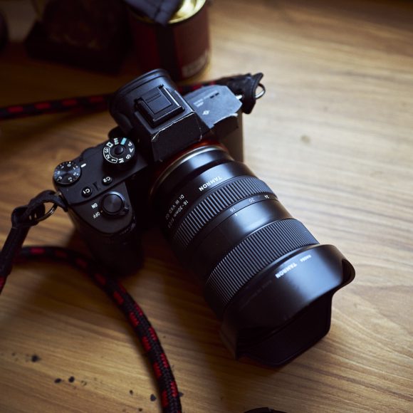 A black digital camera with a large lens rests on a wooden desk near books and a cup.