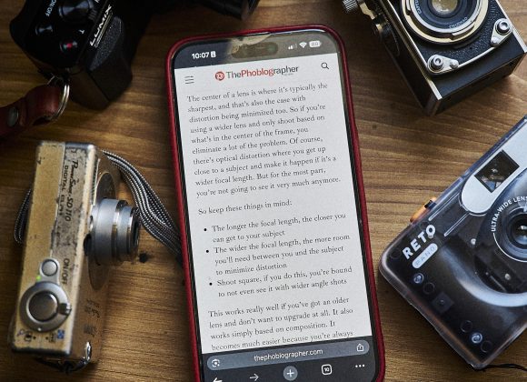 A smartphone displays an article, surrounded by four vintage cameras on a wooden surface.