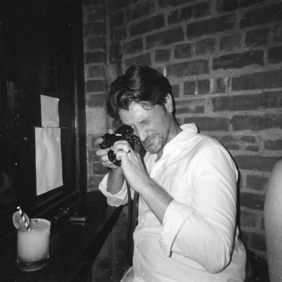 Man in white shirt takes a photo with a camera in a dimly lit bar, next to a drink and a brick wall.