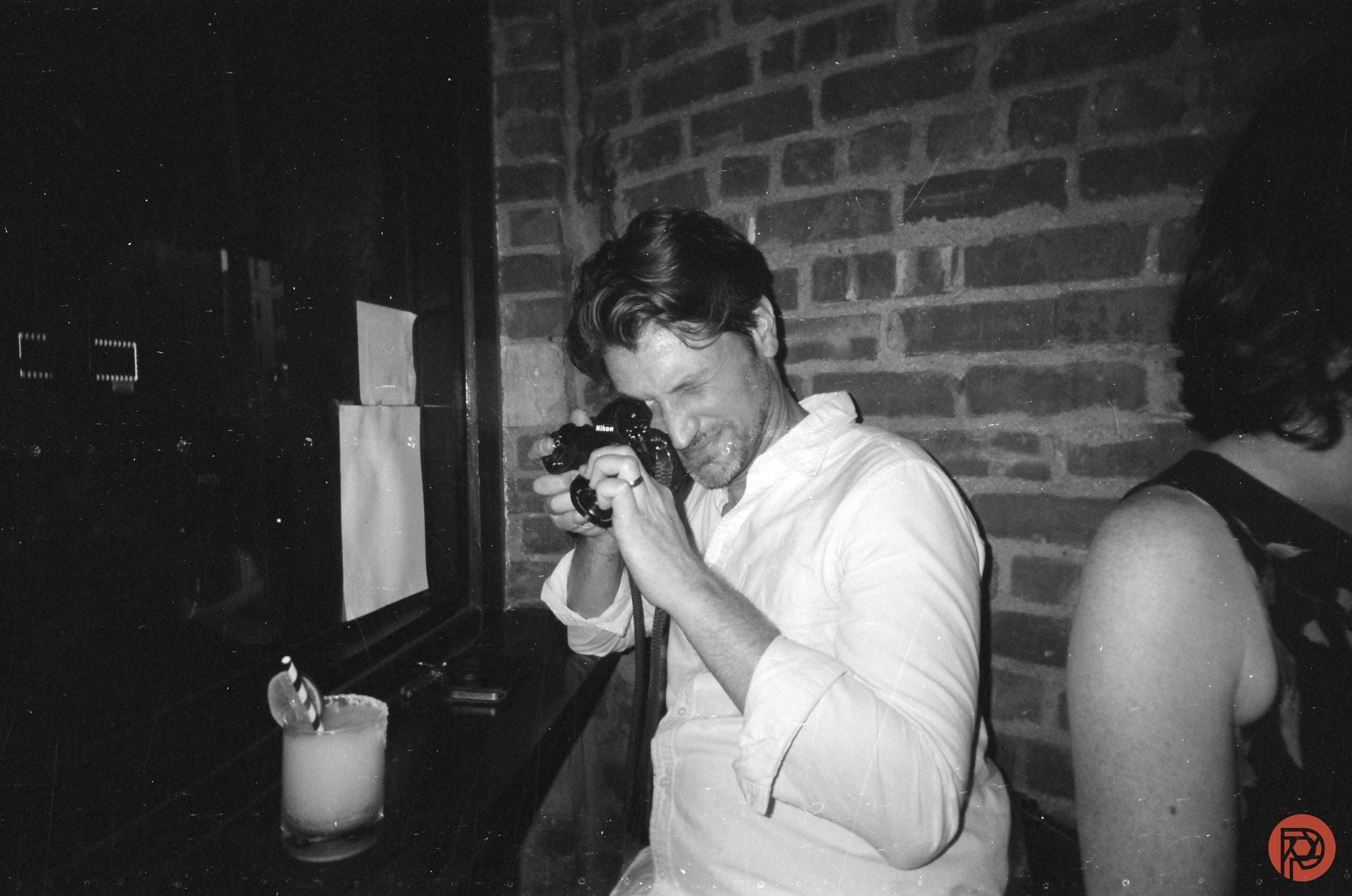 This Free Film Scanning Tool is What Analog Photographers Have Been Waiting For Man in white shirt takes a photo with a camera in a dimly lit bar, next to a drink and a brick wall.
