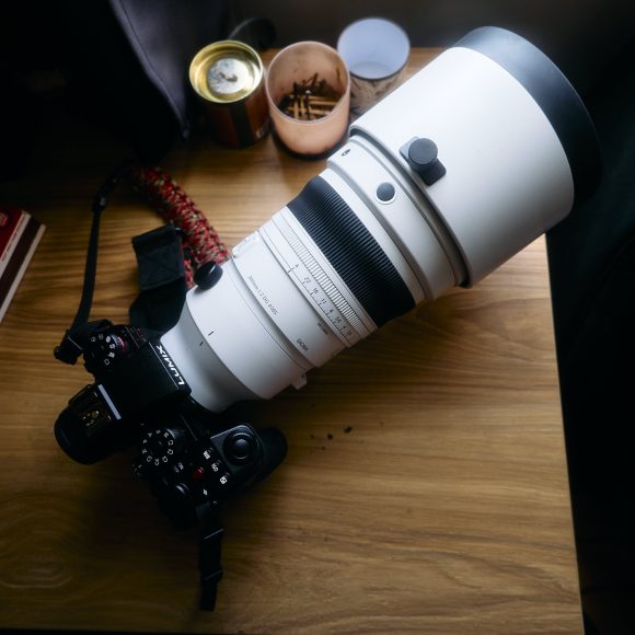 A camera with a large telephoto lens rests on a wooden desk near small containers and a book.