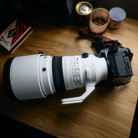 A large white telephoto camera lens attached to a camera body on a wooden desk with various small items.