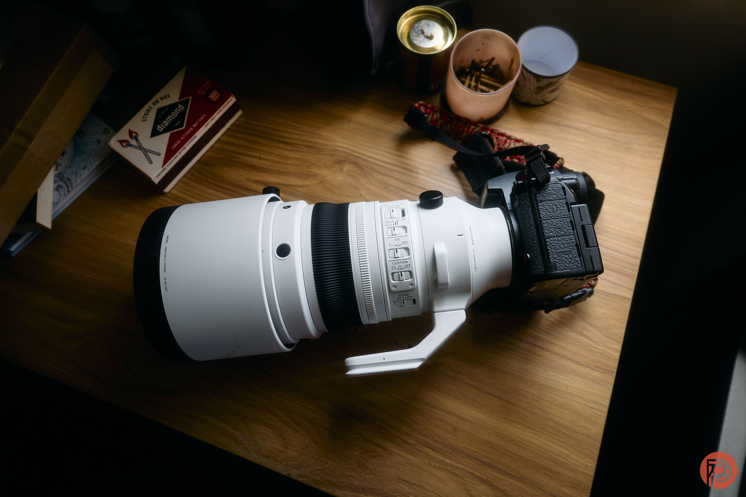 A large white telephoto camera lens attached to a camera body on a wooden desk with various small items.