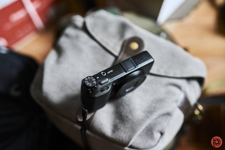 A compact black camera resting on a light gray fabric bag, viewed from above.
