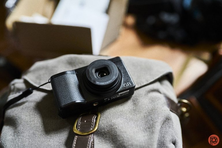 A black compact camera labeled GR rests on a gray fabric bag indoors.