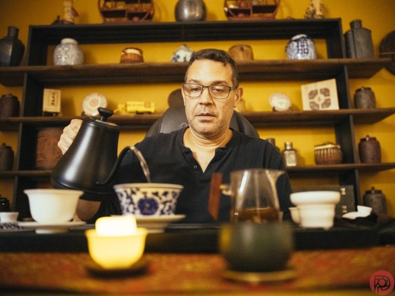 A man pours hot water into a teacup at a table with tea utensils, candles, and shelves behind him.