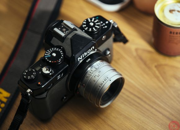 A Nikon Z f camera with a vintage lens on a wooden surface, next to a can and other objects.