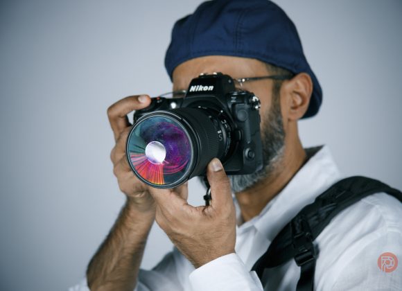 A person in a blue cap and white shirt holding a Nikon camera with a colorful lens, ready to take a photo.