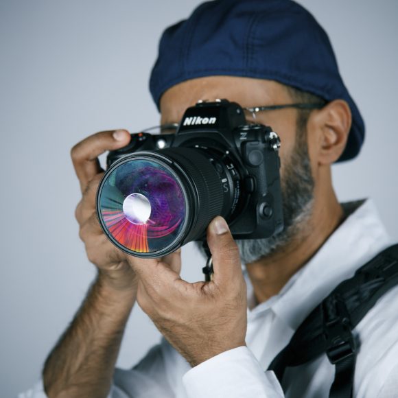 A person in a blue cap and white shirt holding a Nikon camera with a colorful lens, ready to take a photo.