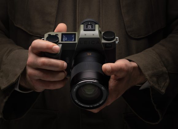 A person in a brown jacket holds a Leica camera with both hands, lens facing forward.