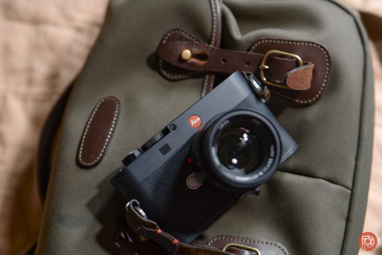 A black Leica camera rests on a green canvas bag with brown leather straps and buckles.