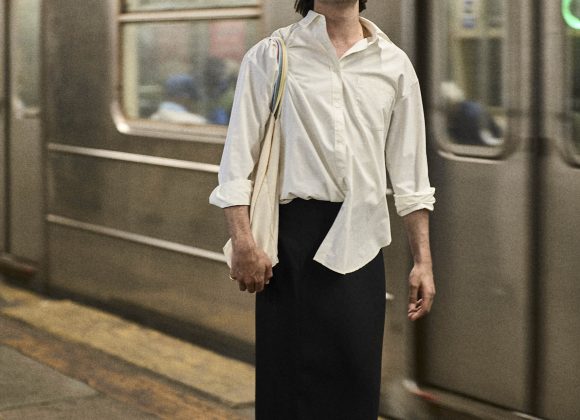 A person in a white shirt and long black skirt stands on a subway platform with a train in the background.