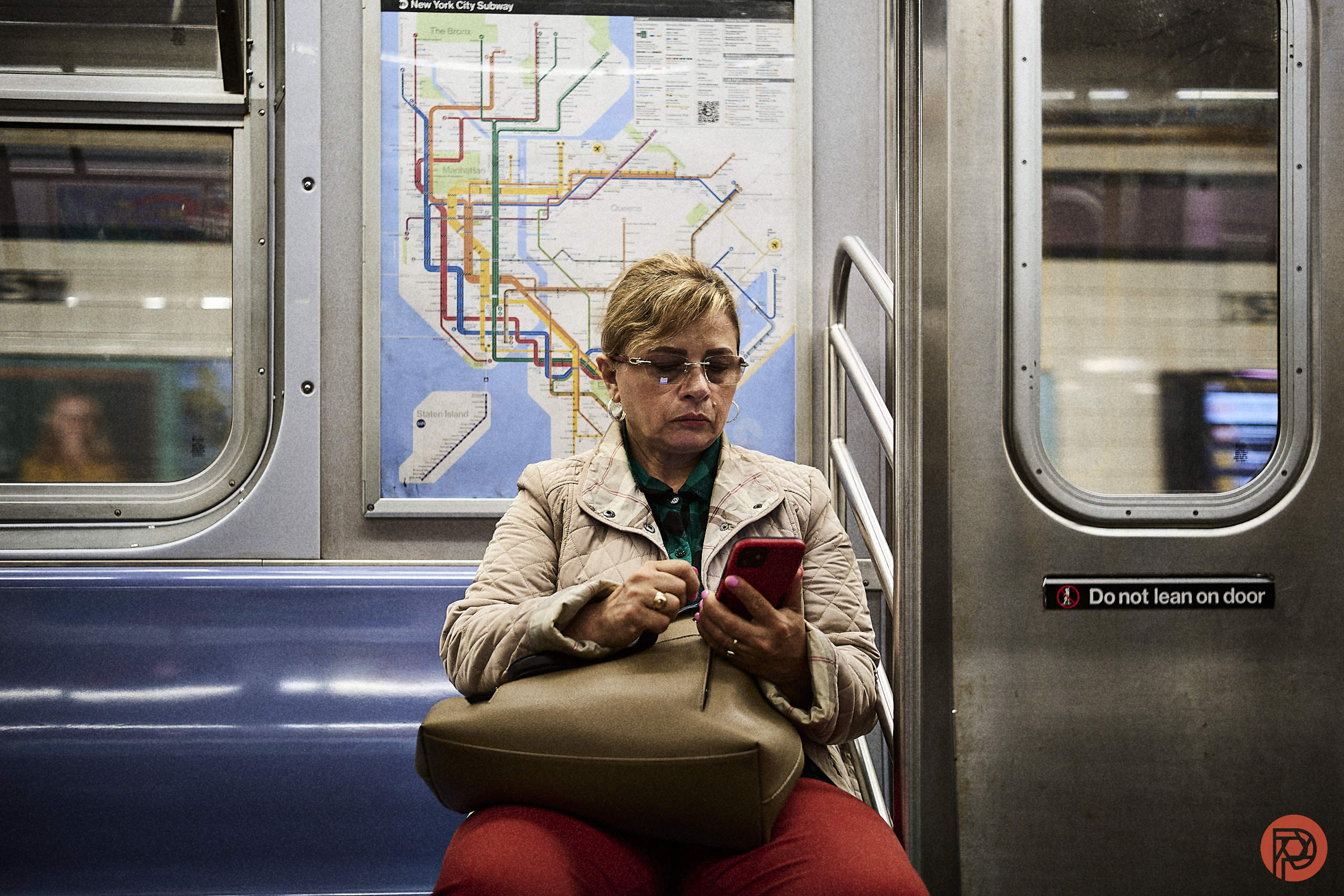 A woman sits on a subway bench, looking at her phone, with a subway map on the wall behind her.