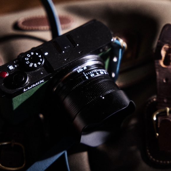 A black Lumix camera with a blue strap rests on a brown leather bag in soft lighting.