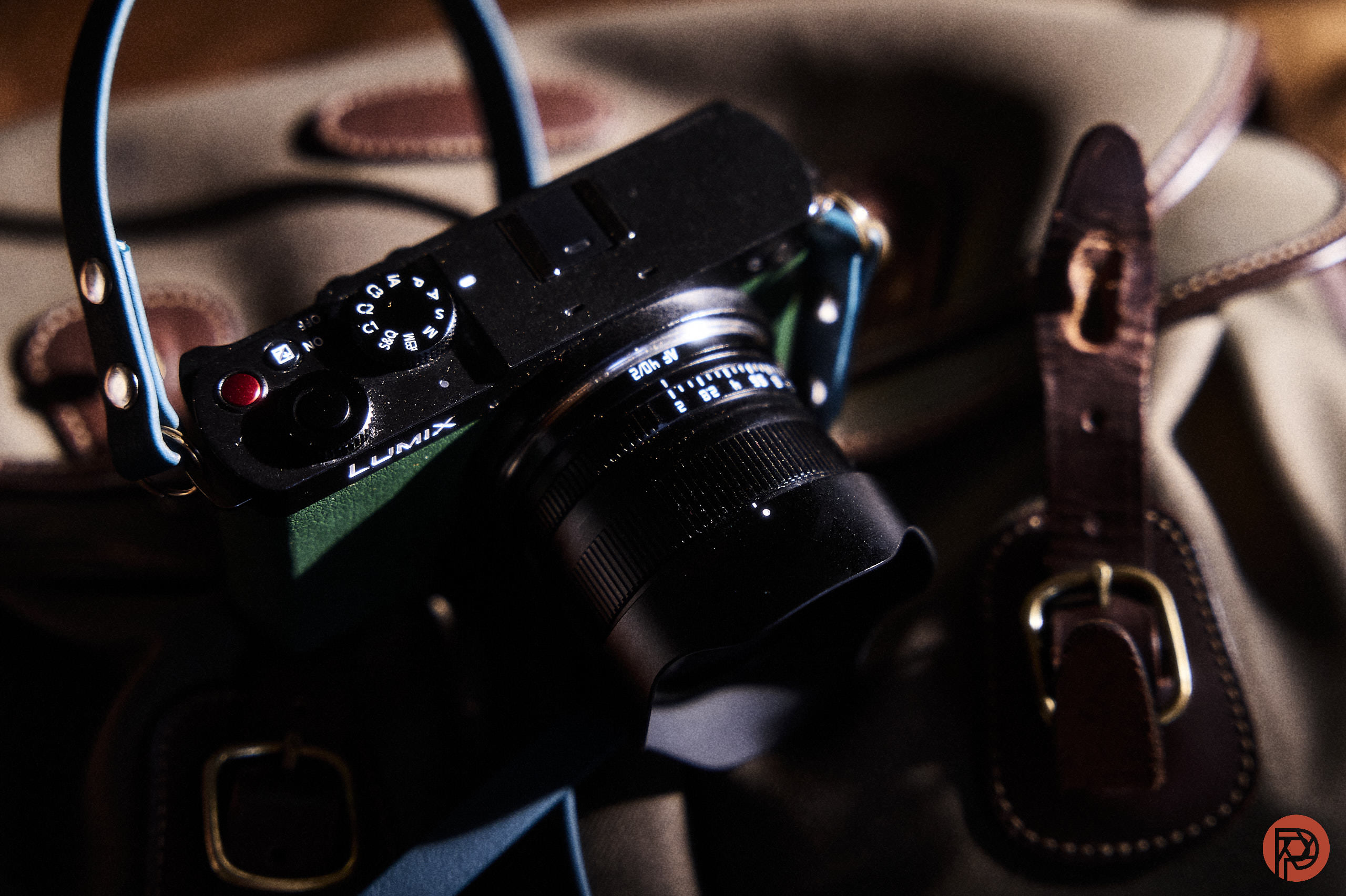A black Lumix camera with a blue strap rests on a brown leather bag in soft lighting.