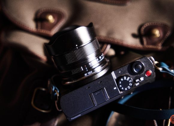 A black digital camera with a wide lens rests on a brown bag with leather accents.