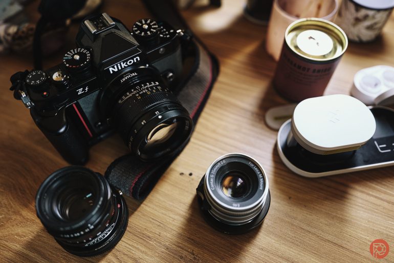 Nikon Z fc camera with three interchangeable lenses on a wooden table, surrounded by various items.