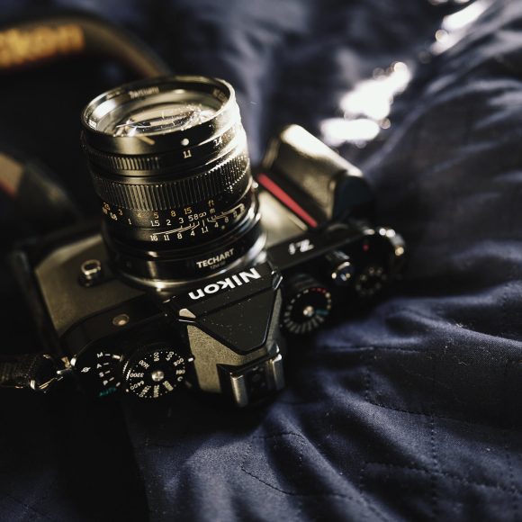 A Nikon camera with a lens rests on a dark quilted fabric with soft, natural lighting.