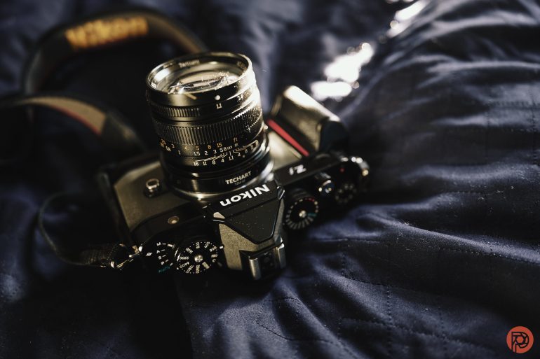 A Nikon camera with a lens rests on a dark quilted fabric with soft, natural lighting.