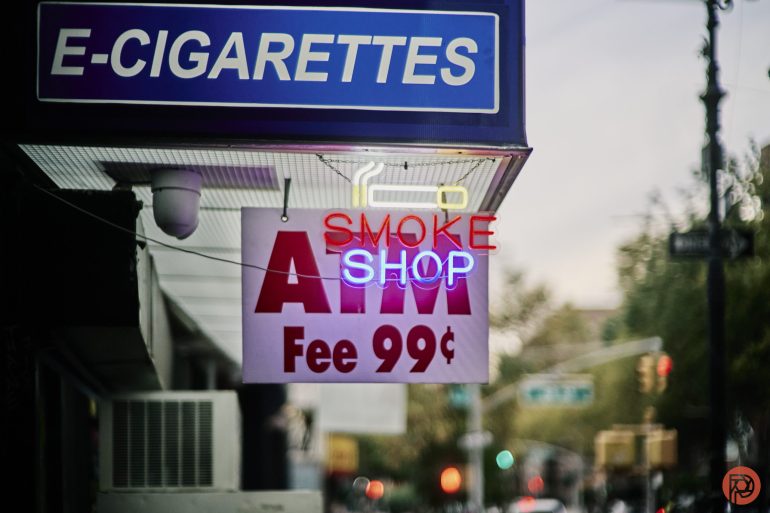 Blue E-CIGARETTES sign above neon SMOKE SHOP and ATM Fee 99¢ signs on a city street storefront.