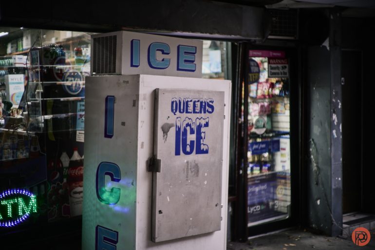 A white ice vending machine labeled QUEENS ICE stands outside a convenience store.
