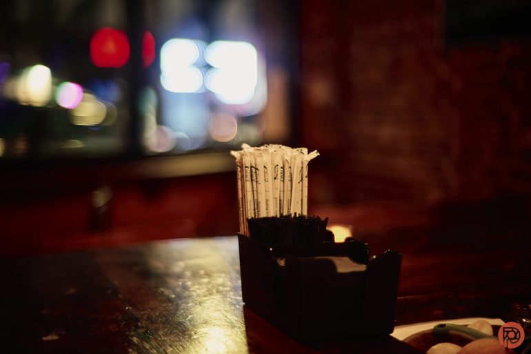 A container of wrapped straws sits on a dimly lit wooden table in a cozy, blurred restaurant setting.