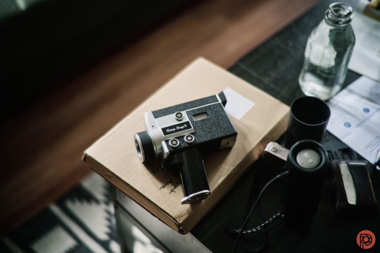 A vintage camera rests on a cardboard box next to a glass bottle and various small items on a table.