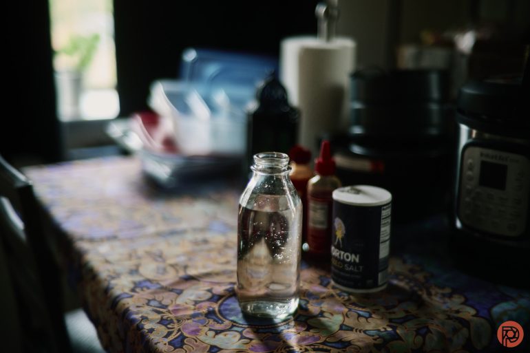 A glass bottle of water on a patterned table with salt, condiments, and kitchen items in the background.