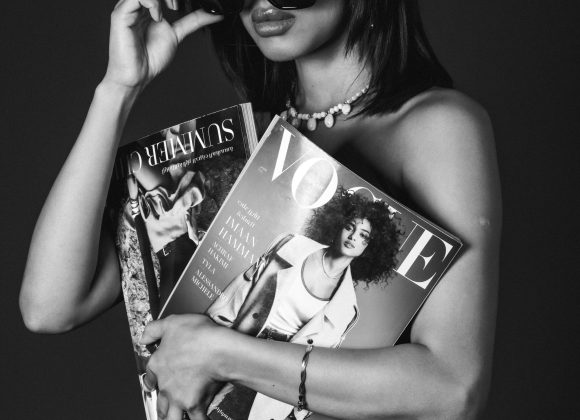 Woman in black dress holds Vogue and Another magazines, lifting sunglasses, with a serious expression. Black and white.
