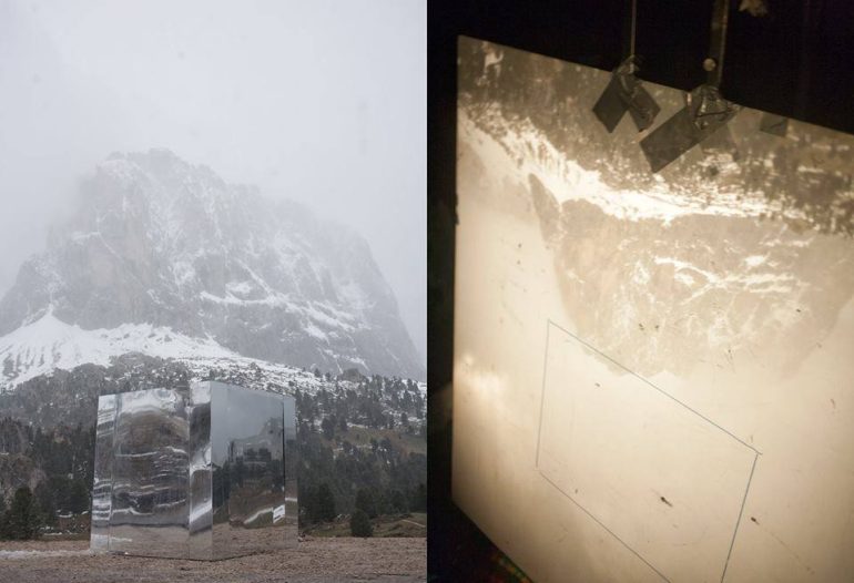 A mirrored cube in a snowy mountain landscape; its reflection projected onto a screen indoors.