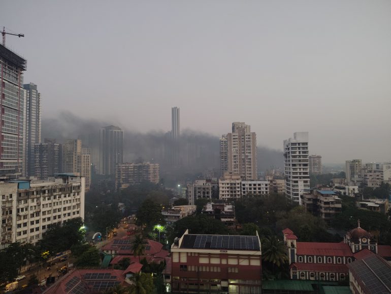 A cityscape at dusk with tall buildings and a large cloud of smoke drifting between them.