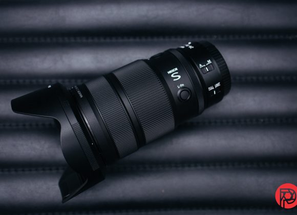 A black camera zoom lens with a hood rests on a dark, ridged surface; red logo in the corner.
